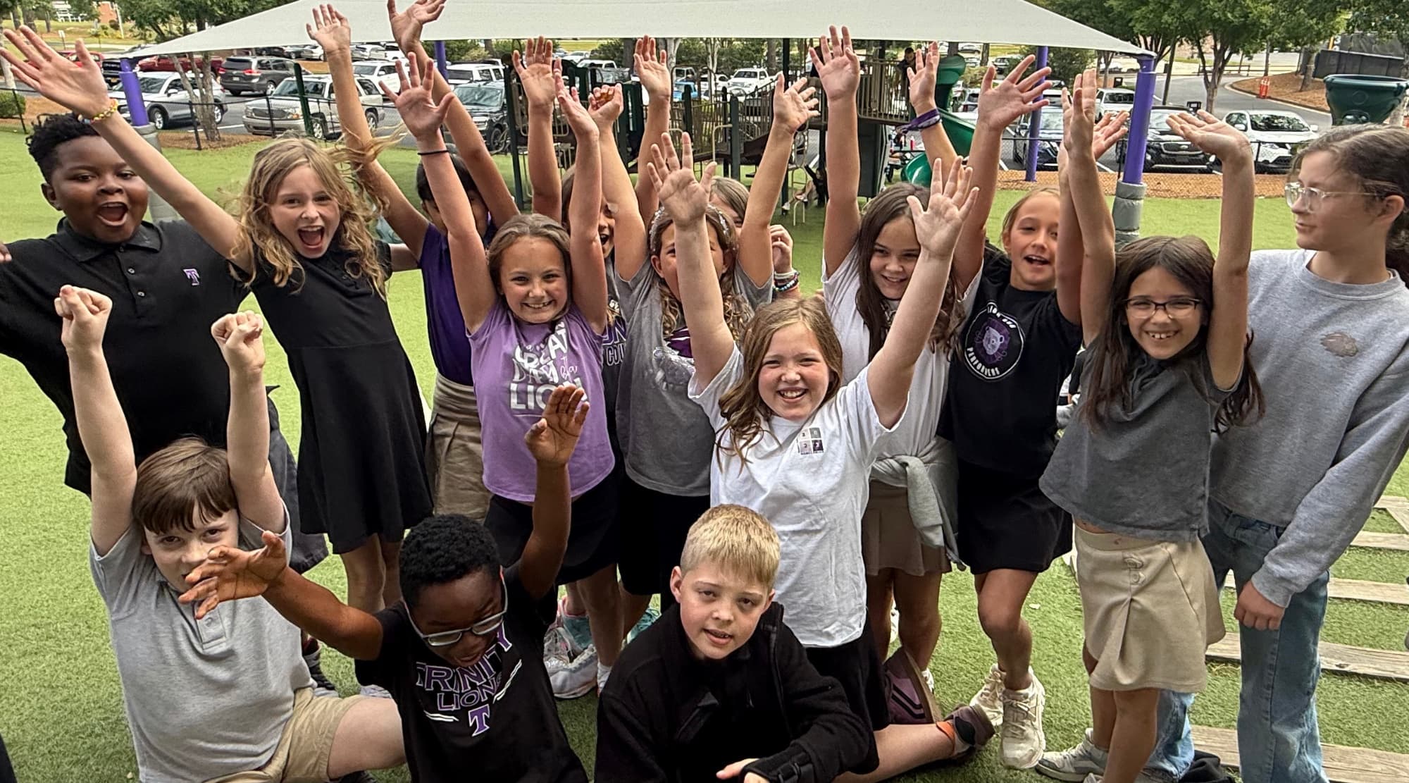 A joyful group of TCS students raising their hands beneath a playground shade